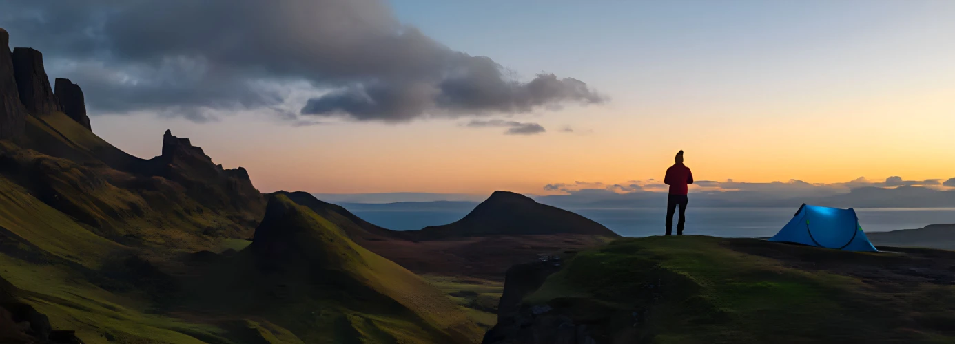 Silhouette of person standing on cliff edge with tent at sunset