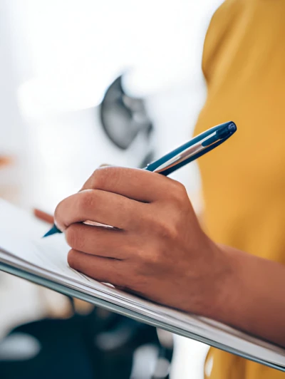 Close-up of a hand holding a blue pen, writing on a clipboard or notepad, with a person wearing a yellow shirt in the background.