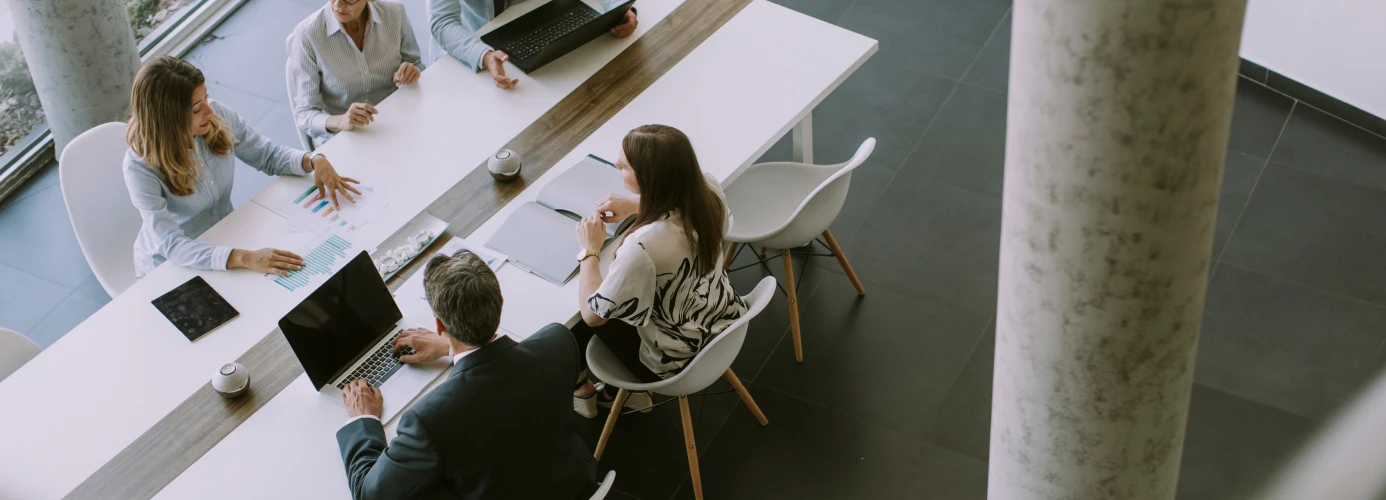 Overhead view of a business meeting with five people seated around a white table, using laptops and discussing documents.
