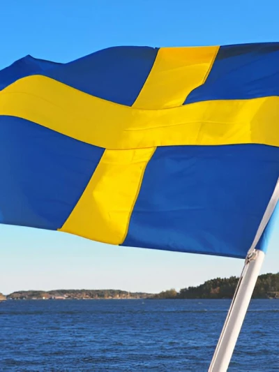 Swedish flag waving in the foreground with a lake and forested shoreline in the background.
